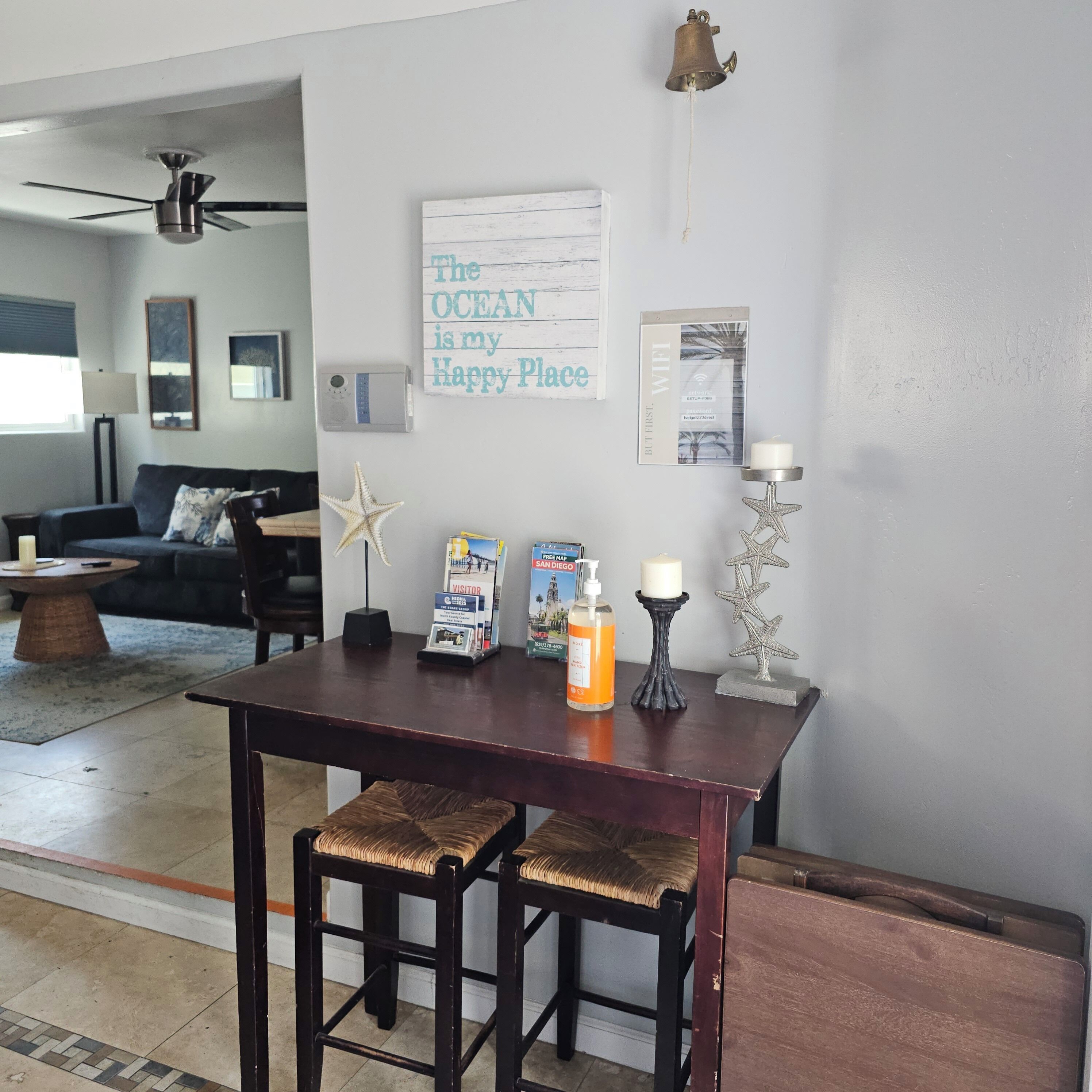 Een woonkamer met een donker houten tafel, twee gestoffeerde krukken, een zeester decoratie, een bordje met de tekst 'The OCEAN is my Happy Place', een kaars en een WiFi-bordje.