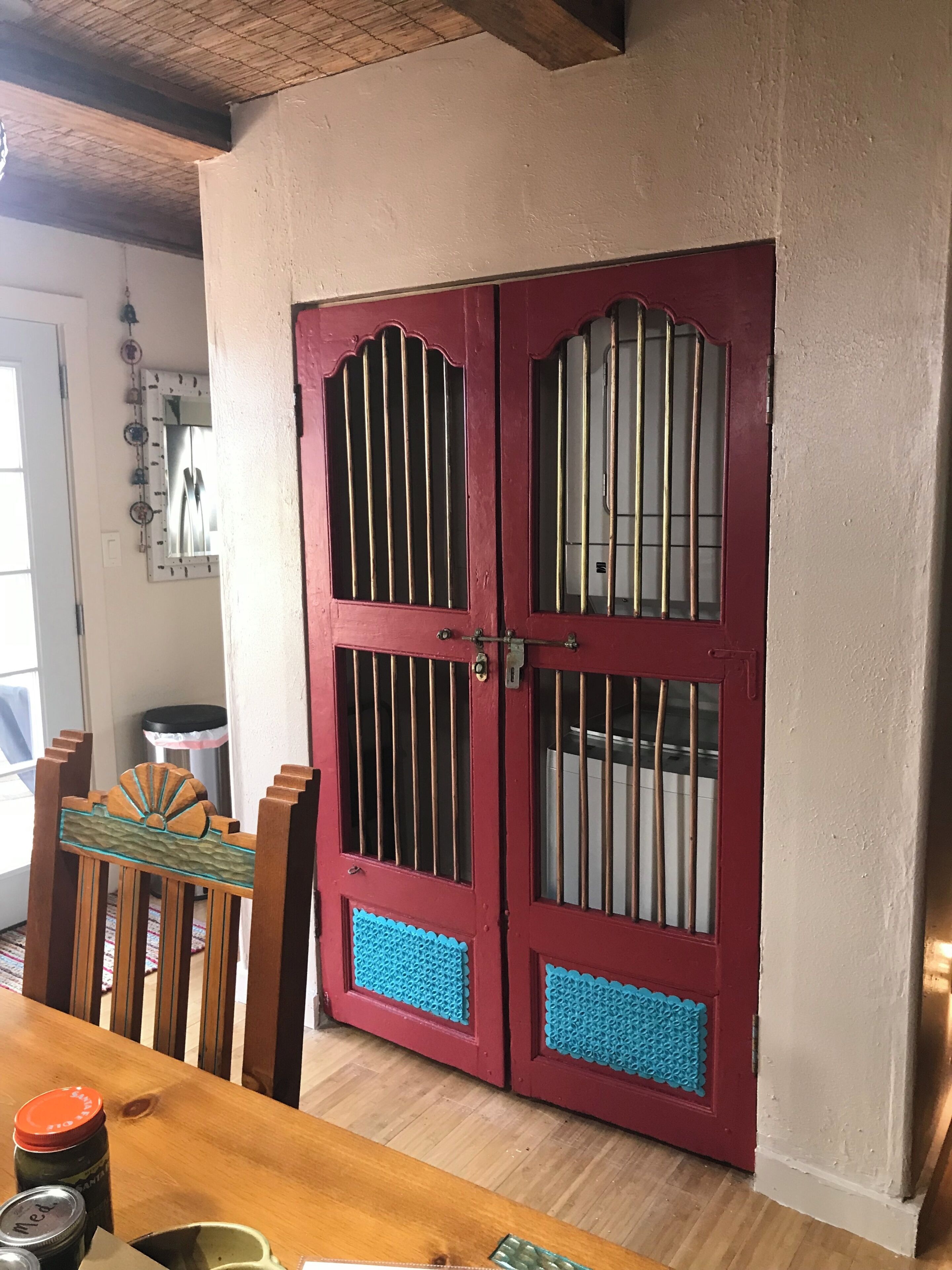 A red door with barred windows, a wooden table with jars, and a patterned mat on the floor.