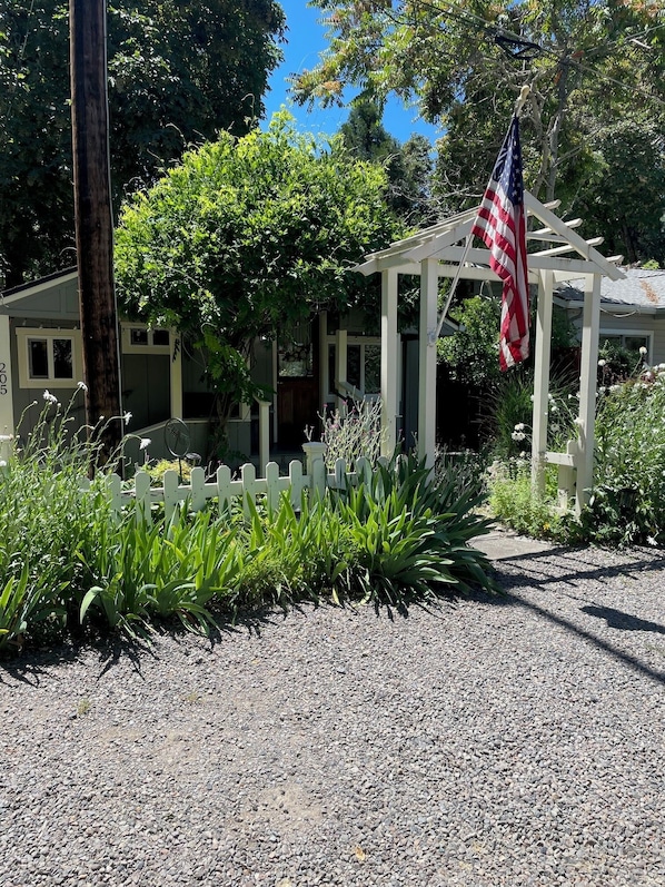 A gazebo with an American flag, surrounded by a white picket fence and lush greenery.