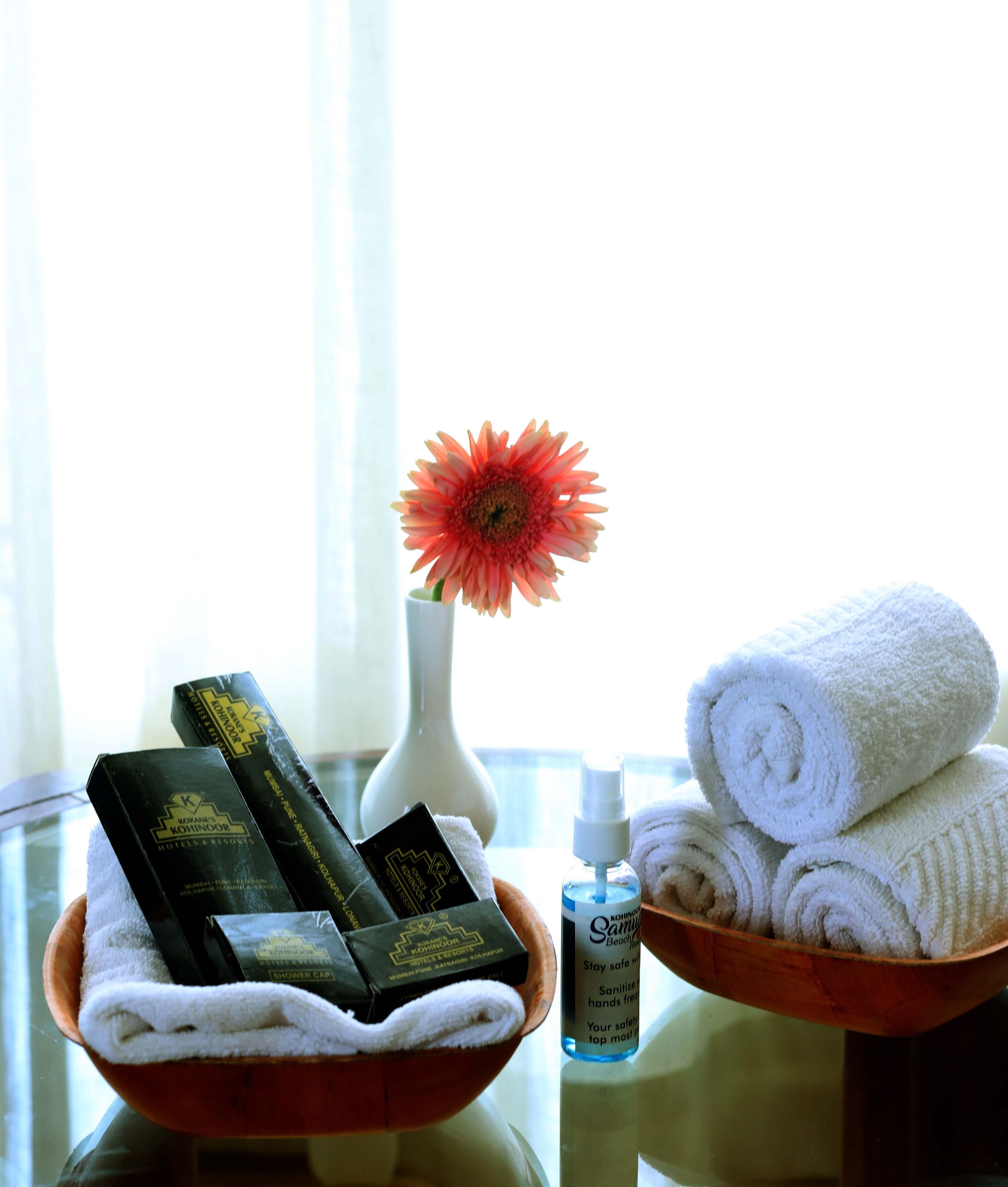 A hotel room with a wooden side table holding a bowl of white towels, a bottle of hand sanitizer, and a white vase with a red flower.