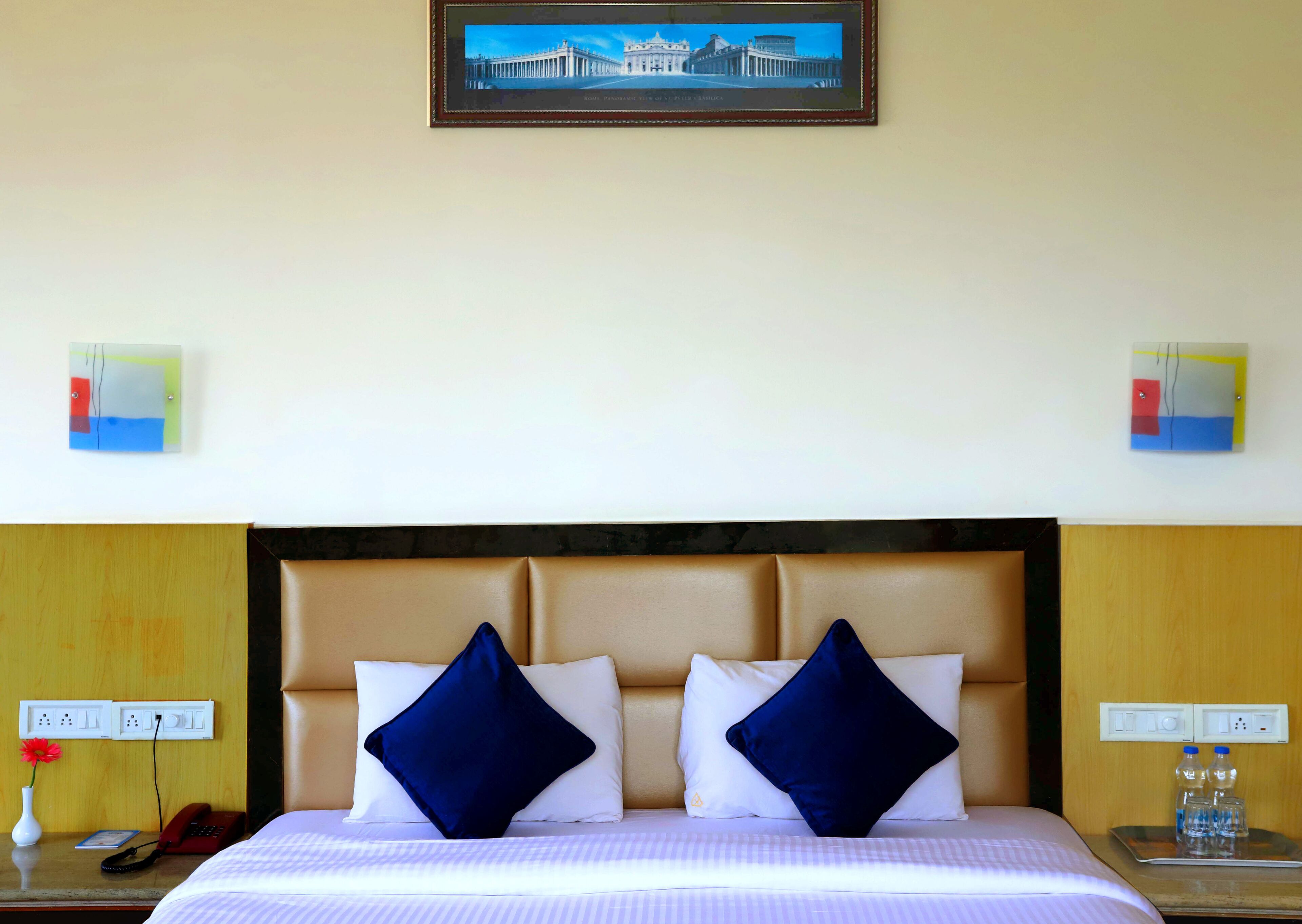 A hotel room with a bed, two pillows, a nightstand, a vase with a flower, a framed picture on the wall, and two wall-mounted light switches.