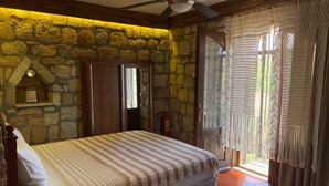 Une chambre avec un mur en pierre, un lit avec une literie à rayures, un ventilateur de plafond et une vue sur l’extérieur, visible à travers une porte vitrée.