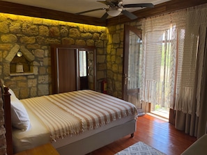 A bedroom with a stone wall, a bed with striped bedding, a ceiling fan, and a view through a glass door.
