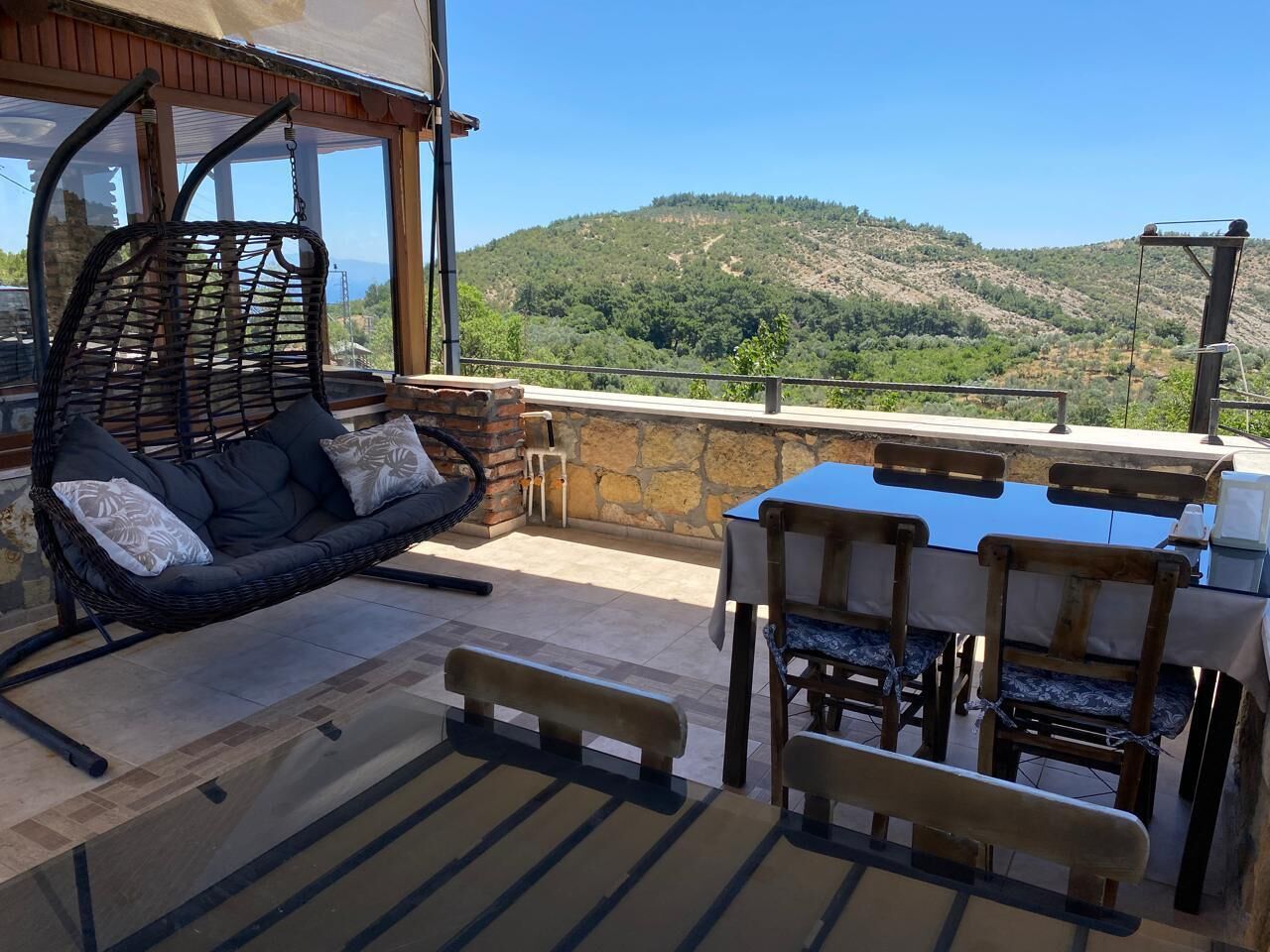 A patio with a wicker sofa, a table with chairs, and a view of a hilly landscape.