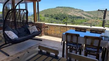 A patio with a wicker sofa, a table with chairs, and a view of a hilly landscape.