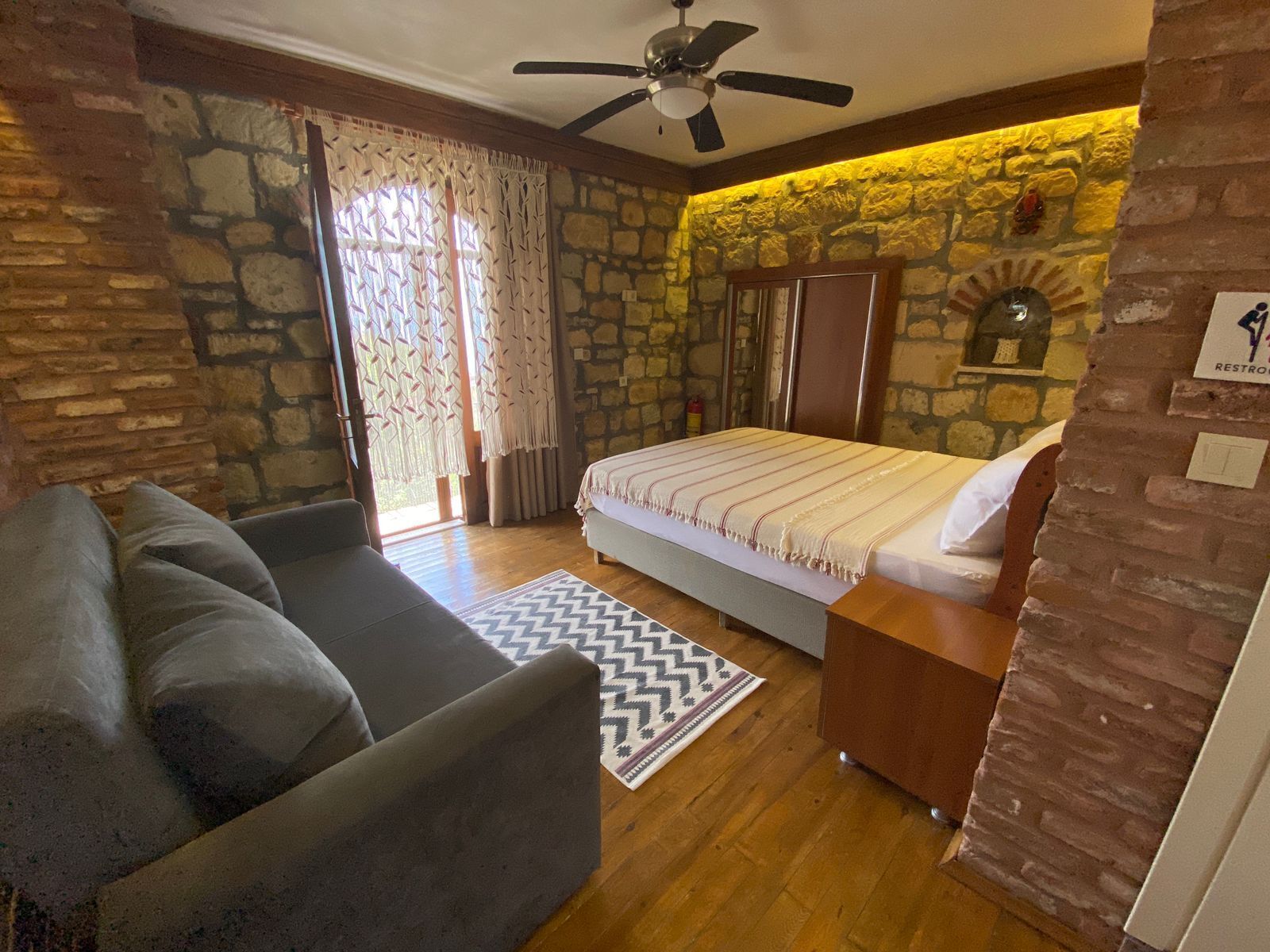 Un dormitorio con cama, sofá, ventilador de techo, paredes de piedra, una ventana con cortinas y un letrero que indica un baño.