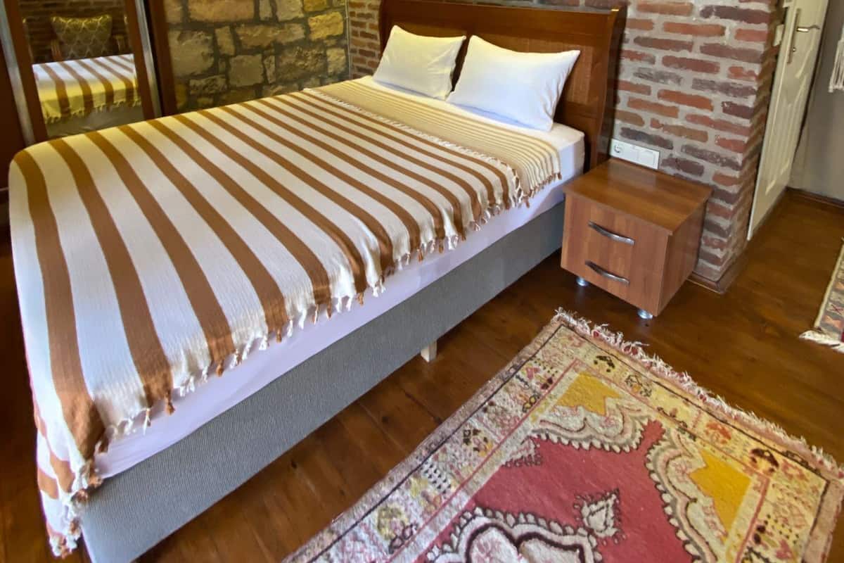 Un letto con una coperta a righe, un comodino, un tappeto decorato, una parete in pietra e una ventola a soffitto.