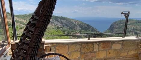 A hanging chair with a cushioned seat and back, positioned on a balcony with a stone wall and a view of the sea and hills.