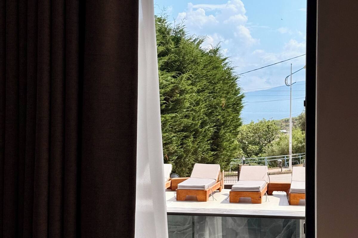 A room with a large window, a curtain partially drawn, and a view of outdoor lounge chairs and greenery.