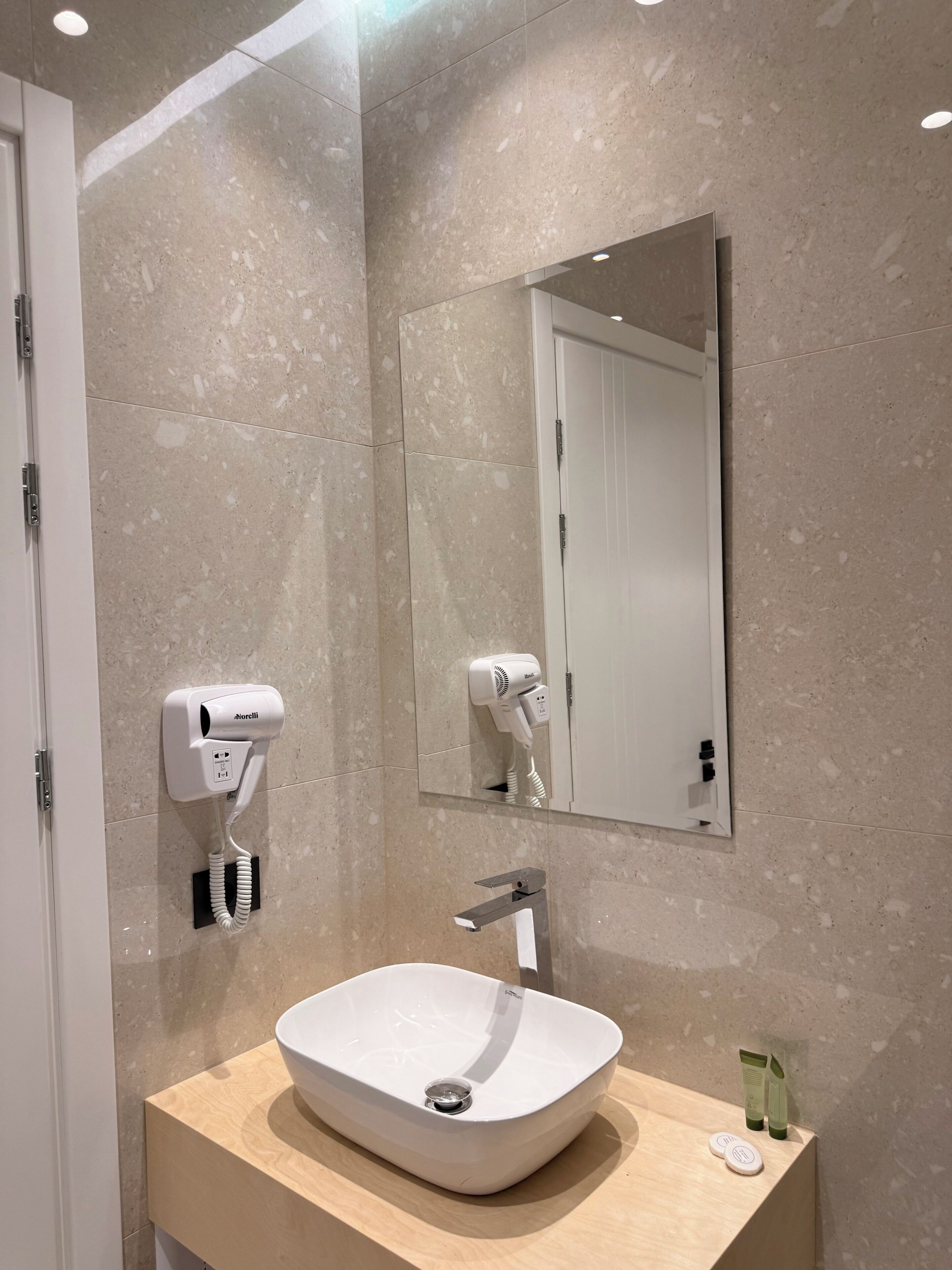 Ett modernt badrum med en beige marmorvägg, en vit tvättställsskål, en handhållen hårtork och en spegel ovanför tvättstället.