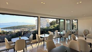 A modern dining area with large windows, a view of the sea, and a cityscape.
