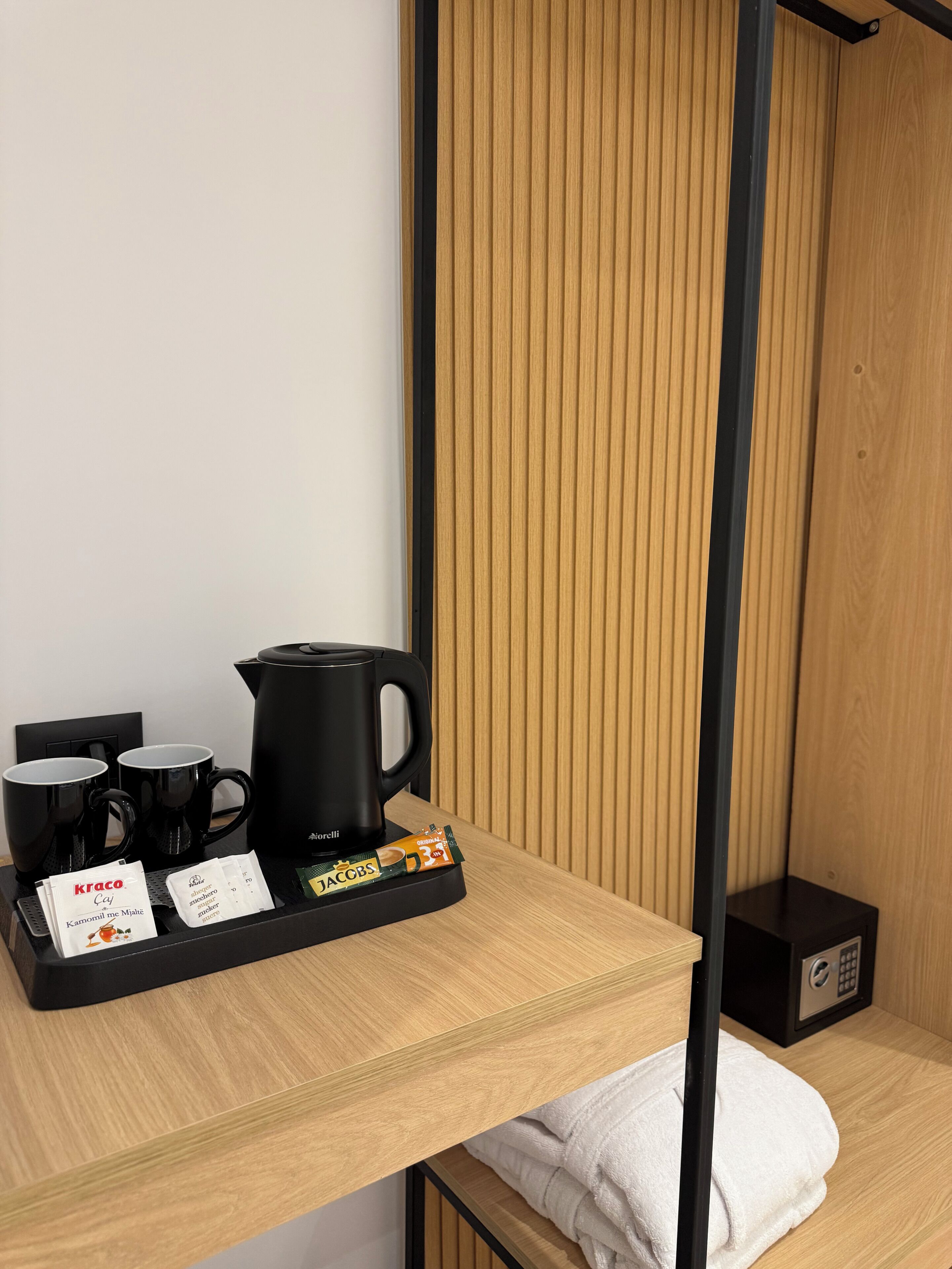 Hotelkamer met een wand van houten panelen, een zwarte koffiezetapparaat, twee zwarte mokken, een dienblad met koffiecups en een witte handdoek op een houten plank.