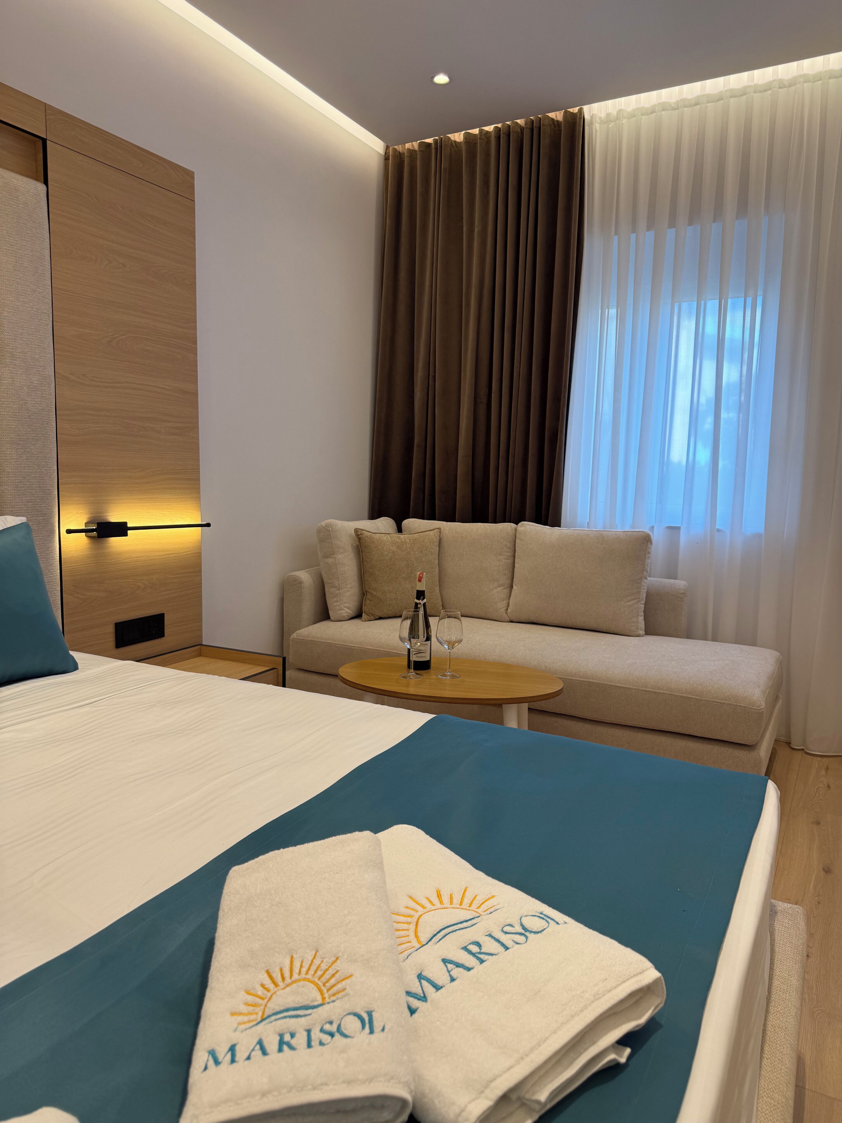 A hotel room with a bed, a sofa, a small table with a bottle and glasses, and a window with curtains.