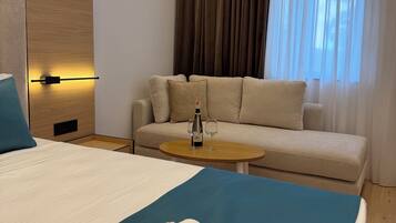 A hotel room with a bed, a sofa, a small table with a bottle and glasses, and a window with curtains.