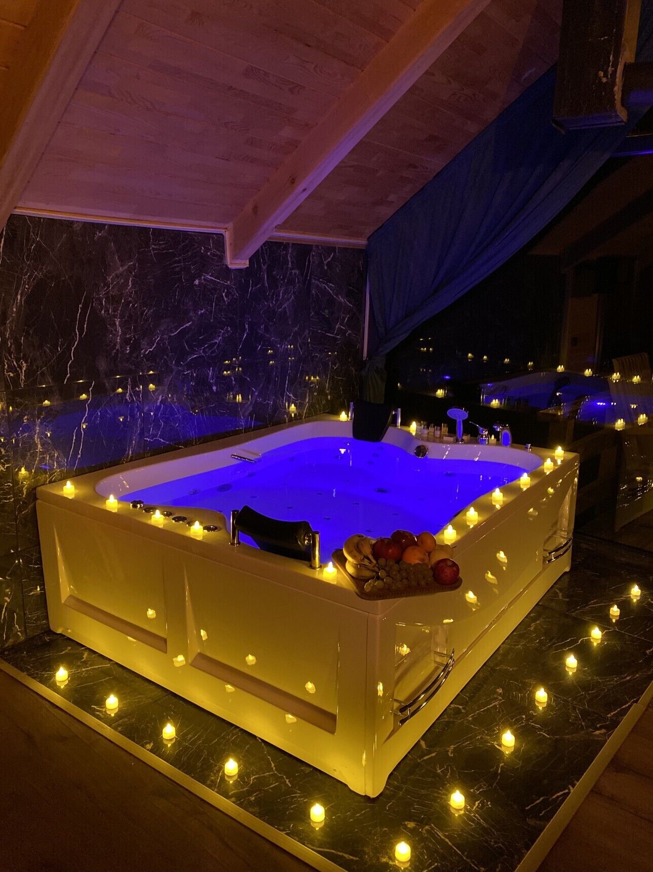 Una bañera de hidromasaje rectangular con luz azul, rodeada de velas y un cuenco de frutas en el borde.