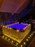 Una bañera de hidromasaje rectangular con luz azul, rodeada de velas y un cuenco de frutas en el borde.