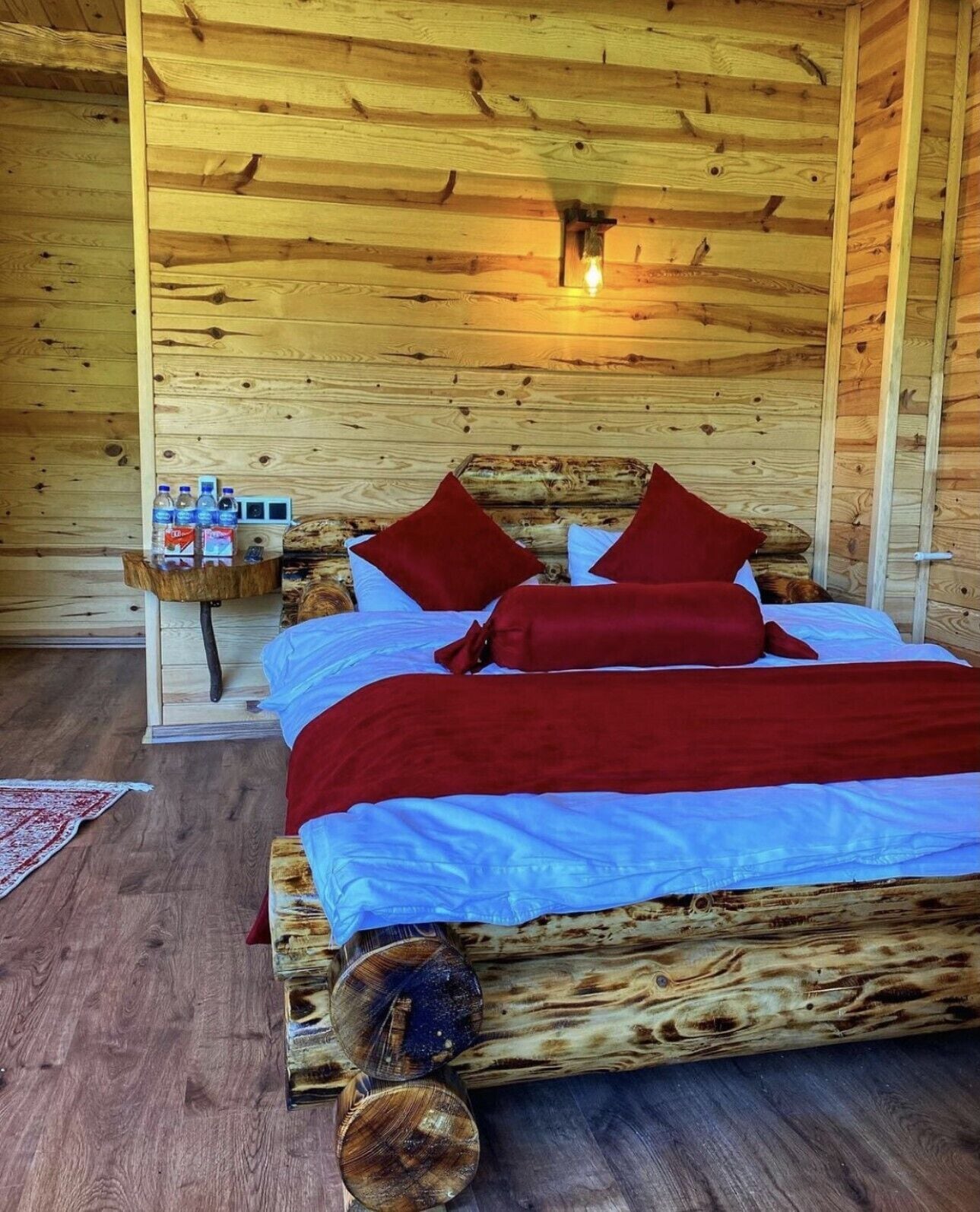 A wooden cabin bedroom with a bed, pillows, a nightstand with water bottles, and a rustic wooden floor.