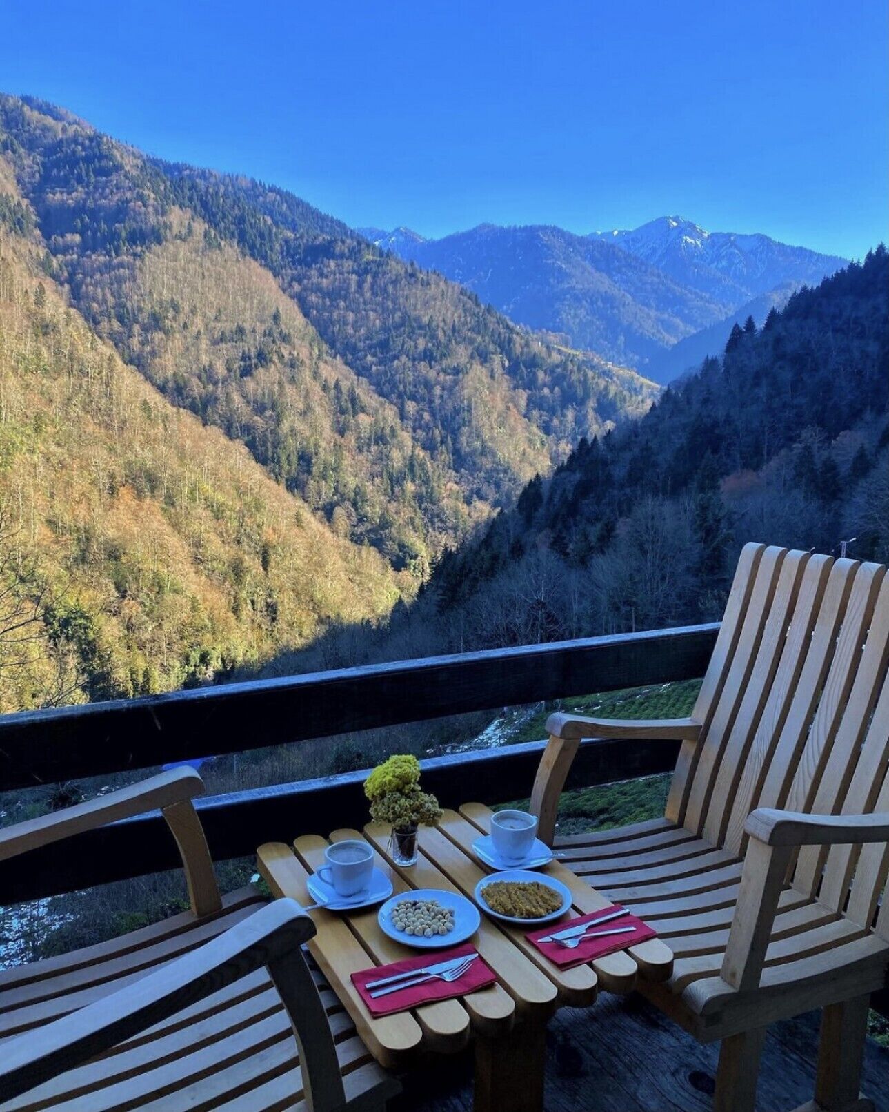 Um deck de madeira com duas cadeiras e uma mesa posta para refeição, com vista para uma paisagem montanhosa.