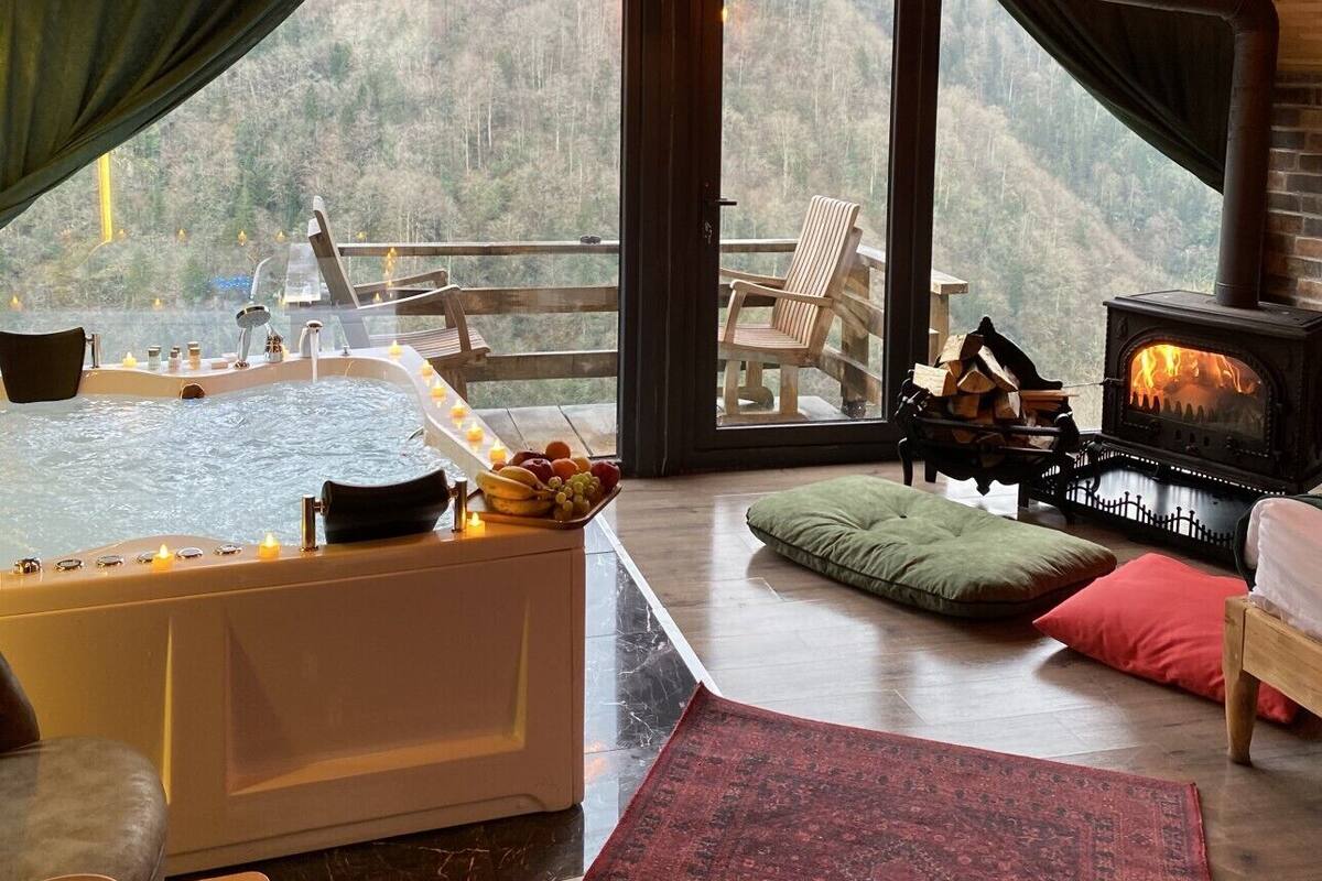 Una cabaña acogedora con jacuzzi, chimenea y un amplio ventanal con vistas a un bosque.