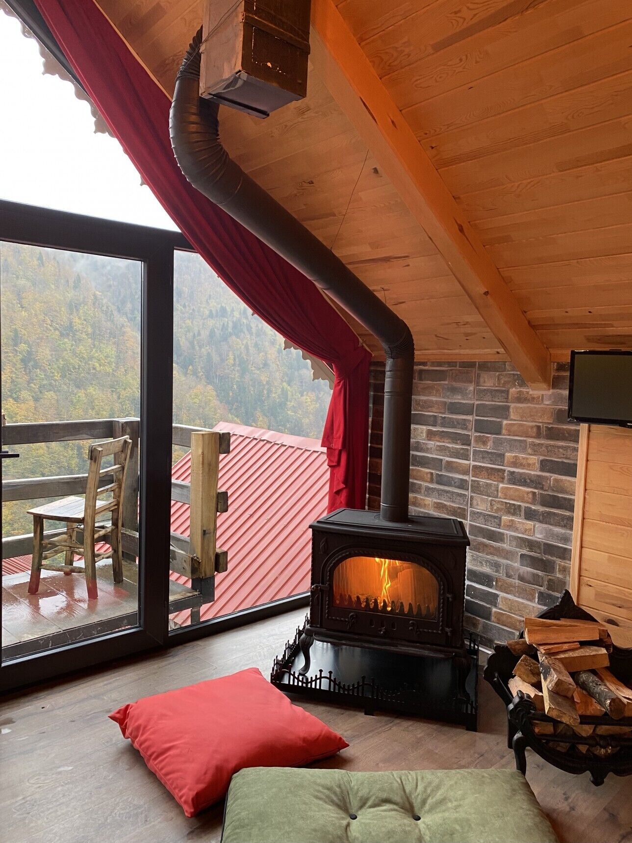 Una habitación acogedora con una estufa de leña, un ventanal con vistas a un edificio de tejados rojos y una silla de madera.