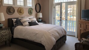 Un dormitorio con cama, dos mesitas de noche con lámparas, un televisor sobre un mueble de madera y una puerta al balcón.