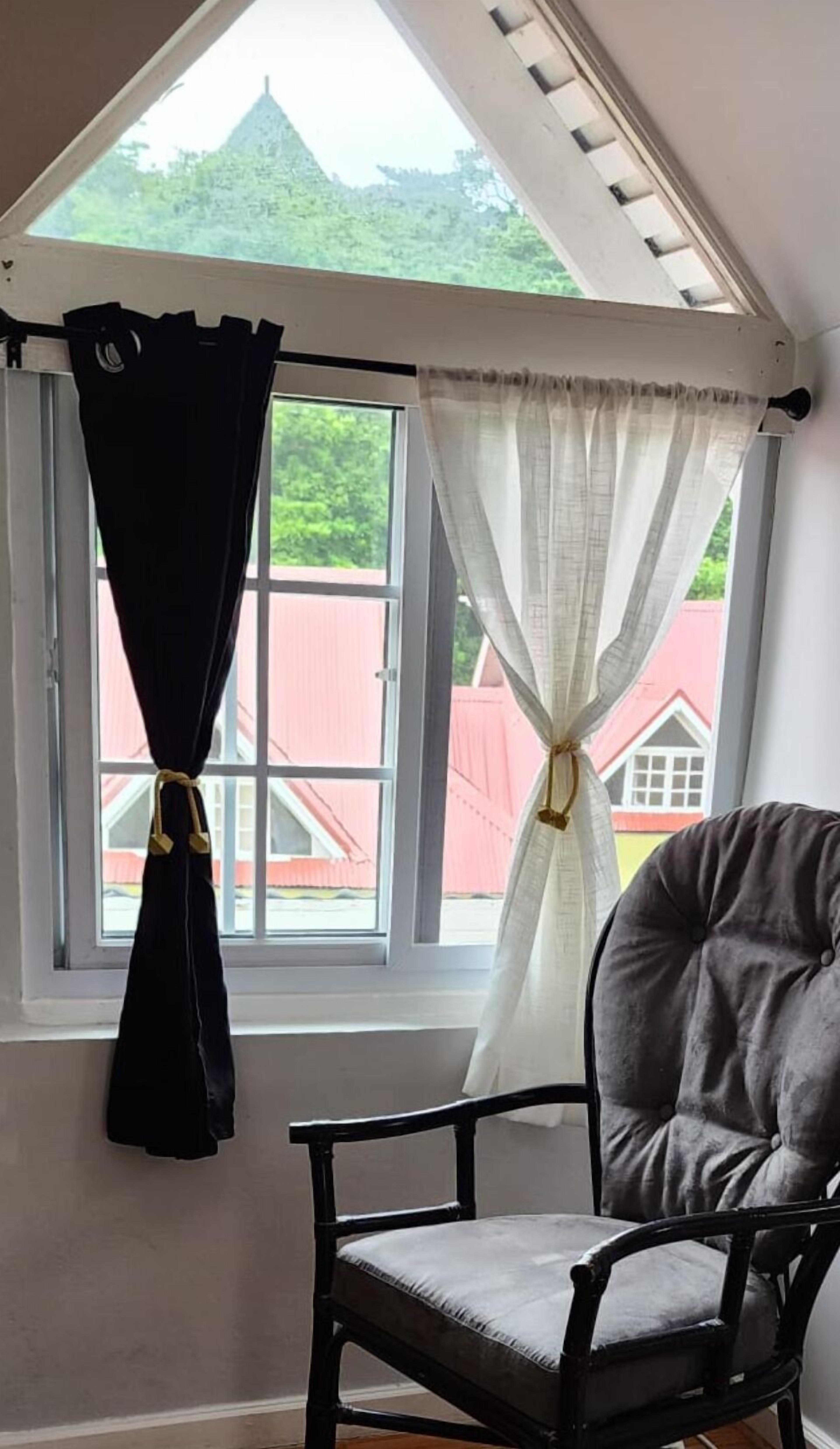 A room with a window, a chair, and a view of a red building outside.