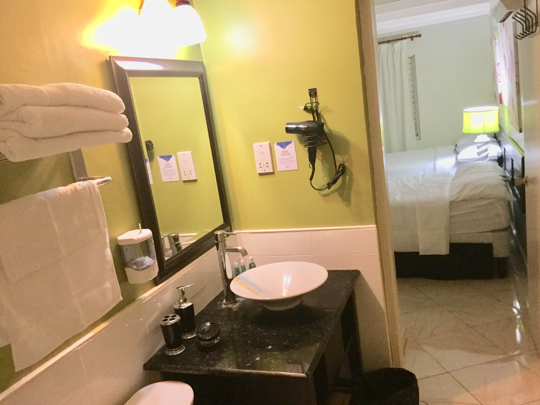 Casa de banho com lavatório, espelho e porta-toalhas. Uma cama visível ao fundo.
