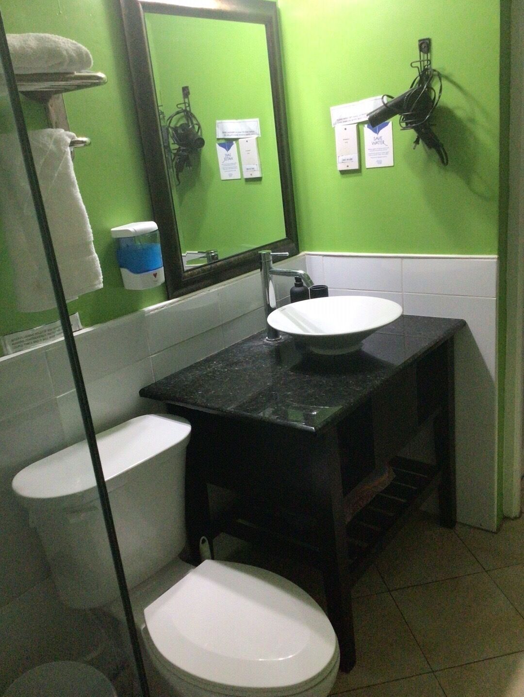 Une salle de bain avec des toilettes blanches, un lavabo et un miroir. Des serviettes sont accrochées au mur, et il y a un distributeur de savon.