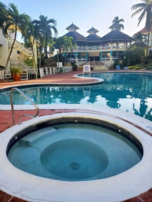 A swimming pool with a hot tub, palm trees, and a covered area with a sign indicating the pool is open. - Mystic Ridge Paradise 2 bedroom Penthouse Condo (Comfy and Relaxing) (Ocho Rios)
