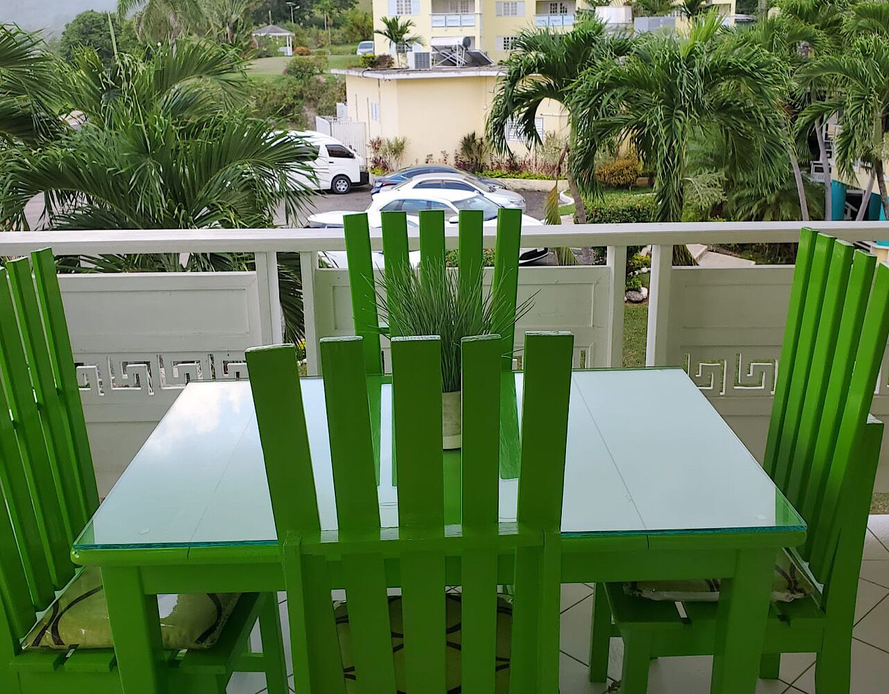 A balcony with green chairs and a glass table, overlooking a street with parked cars and buildings.