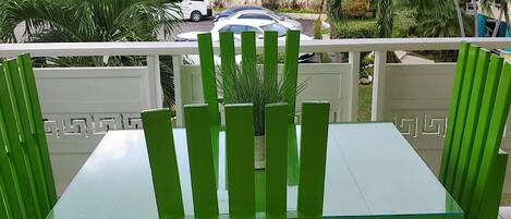 A balcony with green chairs and a glass table, overlooking a street with parked cars and buildings.