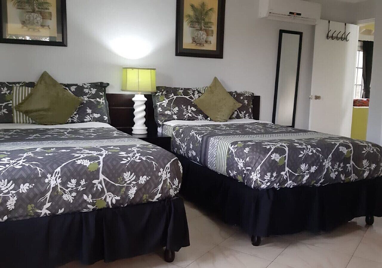 A hotel room with two beds, each with floral-patterned bedding and dark skirts. There is a bedside table with a lamp between the beds.