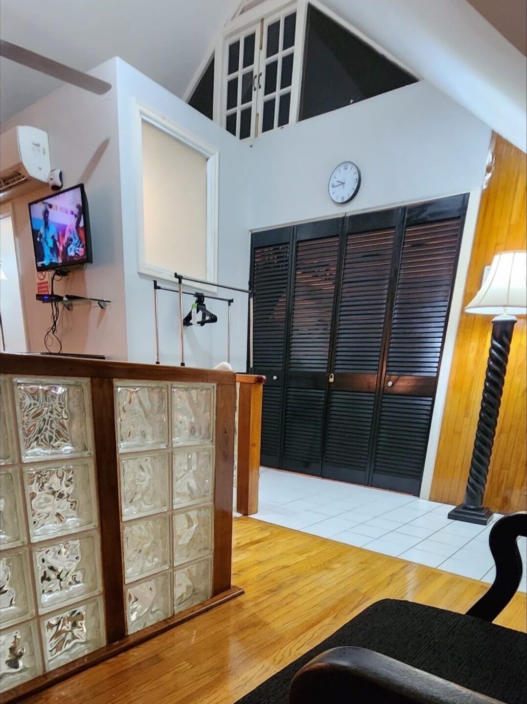 A spacious living area with a wooden floor, a decorative glass partition, a television mounted on the wall, and a large black door with a clock above it.