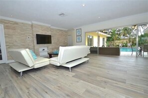 Relax in this stylish living room with a large sofa, flat-screen TV, and sliding glass door leading to a pool and outdoor area.