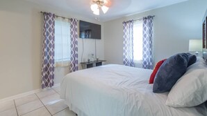 A bedroom with a bed, curtains, a ceiling fan, a flat-screen TV, and a bedside table. - Experience the comfort you deserve while on vacation! (Deltona)