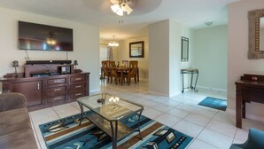 A living room with a TV, entertainment unit, sofa, coffee table, and dining area with chairs and a chandelier. - Experience the comfort you deserve while on vacation! (Deltona)