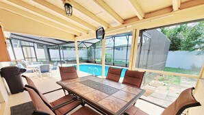 An outdoor patio with a table and chairs, a pool, and a covered area with windows. - Experience the comfort you deserve while on vacation! (Deltona)