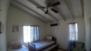 Una habitació amb ventilador de sostre, dos llits, una finestra amb cortines i una cadira.