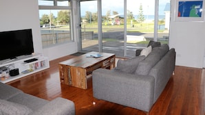 A living room with a large TV, grey sofas, wooden coffee table, and a view of the outdoors through large windows.