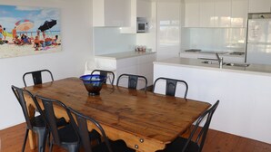 A dining area with a wooden table and chairs, a kitchen with white cabinets and a microwave, and a beach scene painting on the wall.