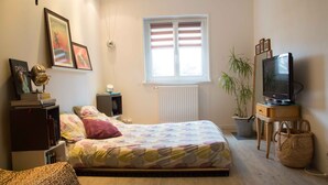 Un dormitorio con cama, televisor, radiador, ventana y estantería.