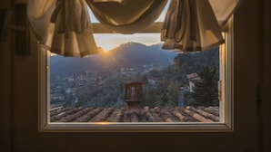 A window with a view of a town and hills, curtains partially drawn, and a chimney on the roof.