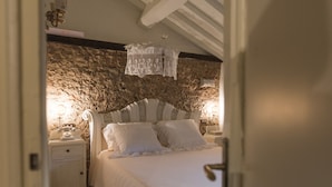 A bedroom with a stone wall, a white bed with ruffled pillows, and a nightstand with a lamp.