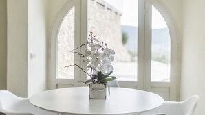 A round white table with a potted orchid in the center, set against a white wall with a large arched window.
