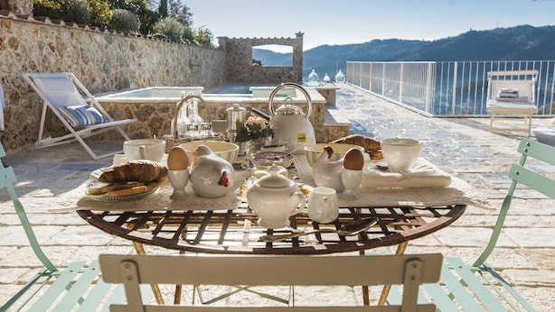 A breakfast setup on a terrace with a stone wall, a pool, and a view of the mountains.