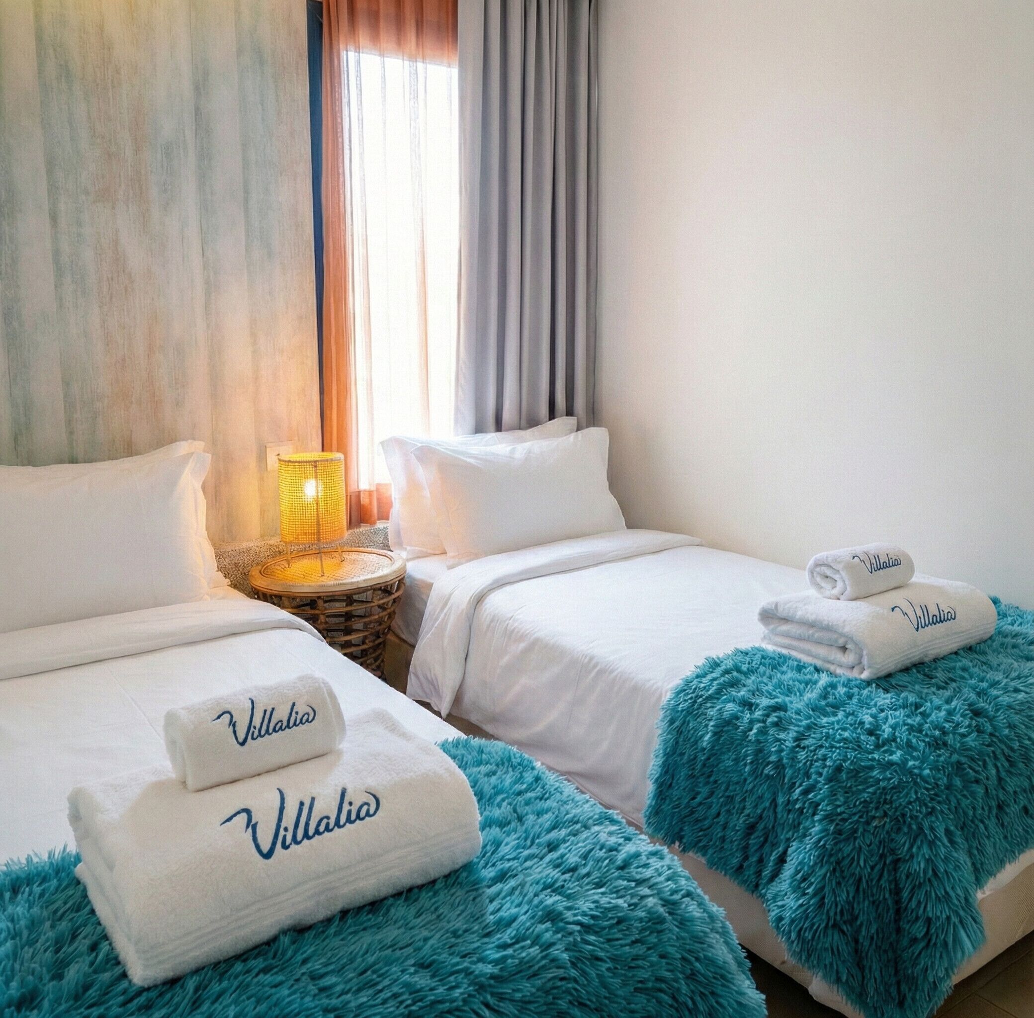 A hotel room with two beds, each with a white bedspread and a blue throw blanket. There are two towels with the name 'Villalba' on them, and a small lamp on a side table.