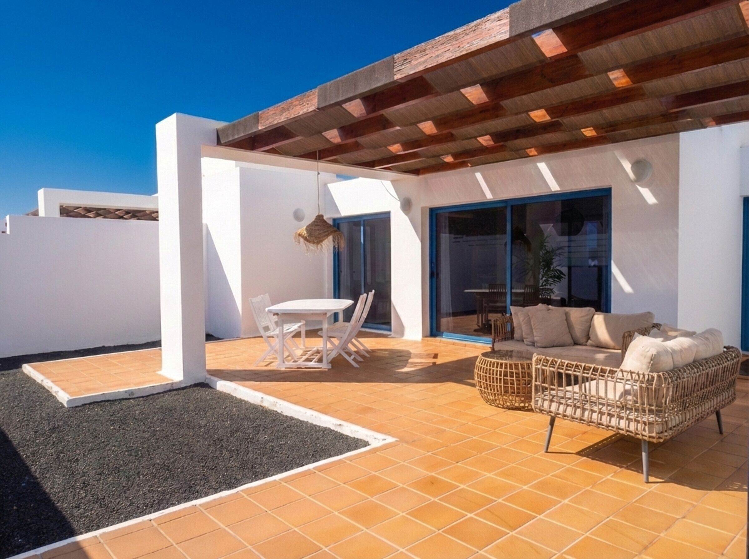 Un patio moderno al aire libre con una mesa y sillas blancas, un sofá de mimbre y una zona de estar cubierta con techo de madera.