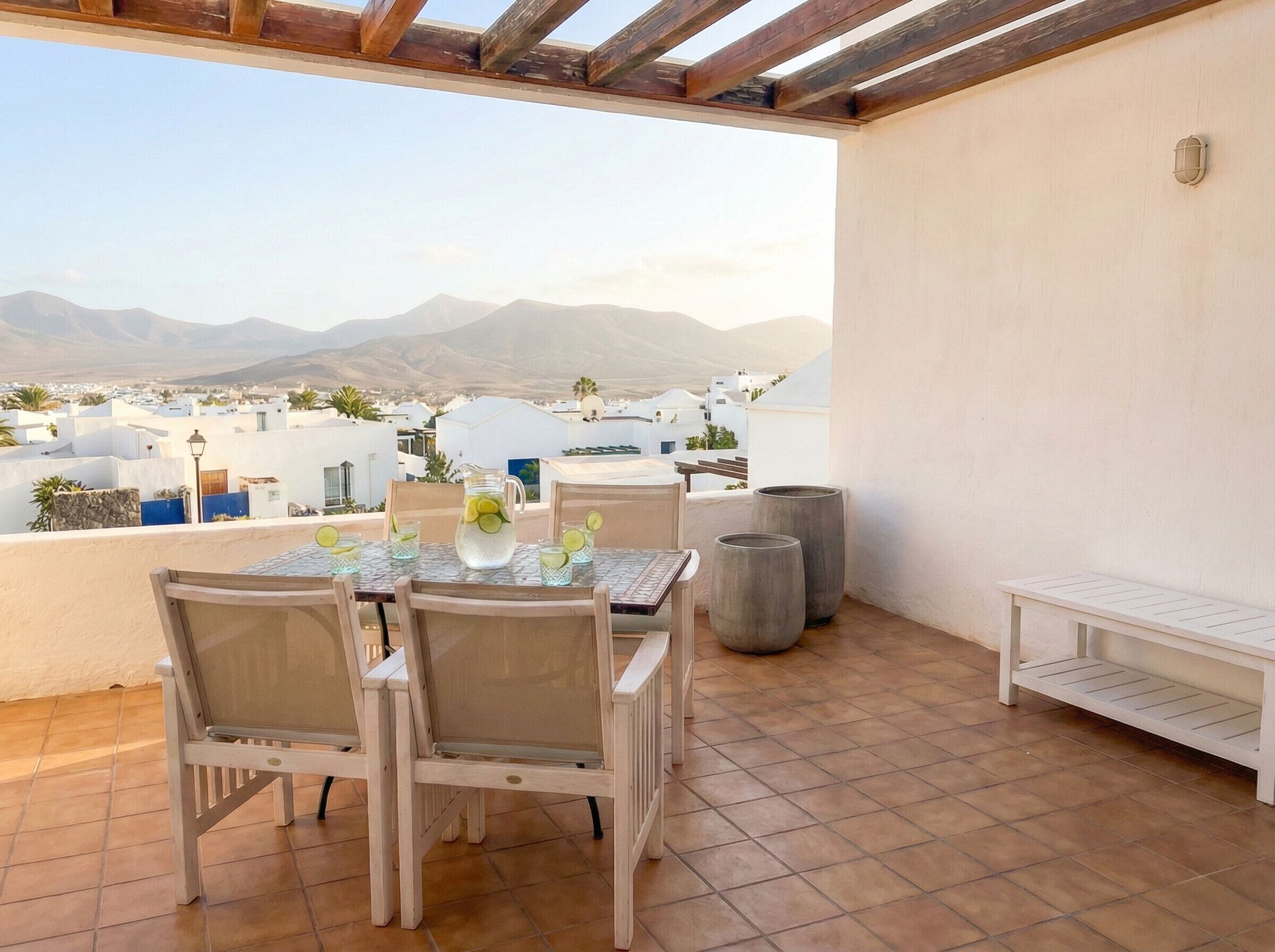 A terrace with a table set for two, chairs, a bench, and a view of a coastal town with mountains in the background.