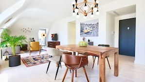 Een ruime loft met een eethoek, bestaande uit een houten tafel en stoelen, een hangende kroonluchter en een grote potplant.