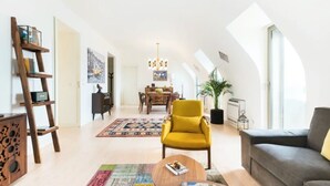 Een ruime loftappartement met hoge plafonds, een eetgedeelte, een woonkamer met een bank en fauteuil, en een groot raam dat natuurlijk licht binnenlaat.